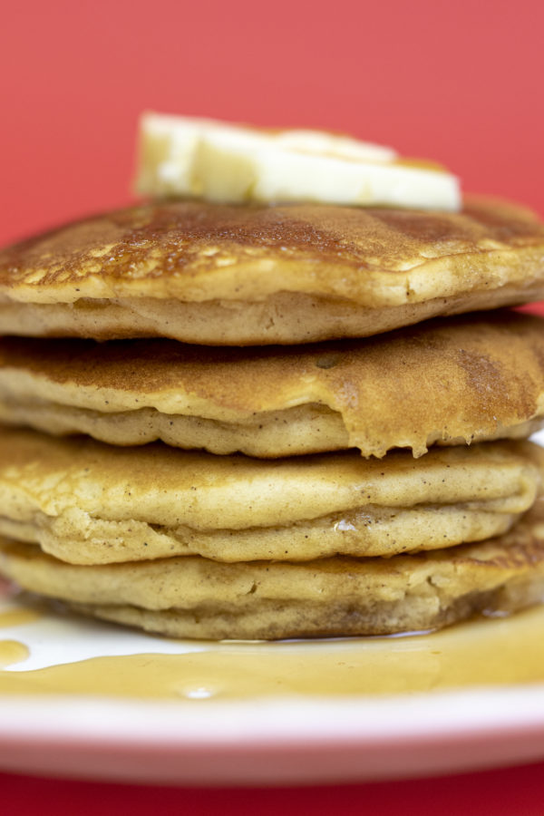 Apple cinnamon pancakes with butter and syrup drizzling over the sides on a white plate with a red backdrop