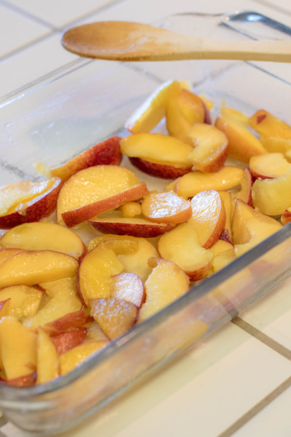 sliced peaches coated in sugar in a baking dish with a wooden spoon to mix it with.