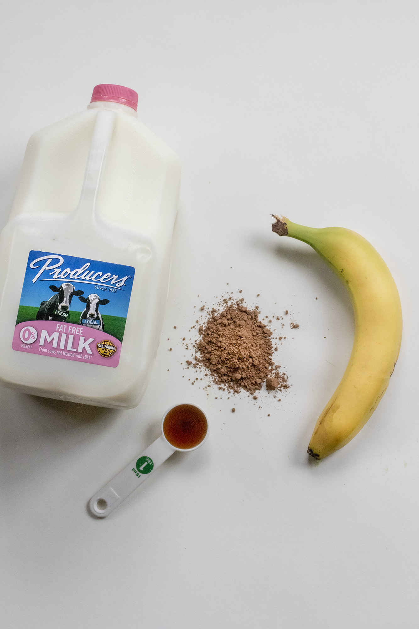 Producers Fat Free Gallon of milk, 1 tablespoon of maple syrup, cinnamon, and a banana laying on a flat white background.